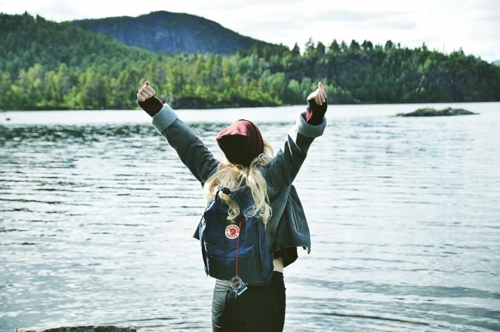A person with long hair, wearing a hooded jacket and backpack, stands with arms raised facing a serene lake. In the background, there are forested hills under a cloudy sky.