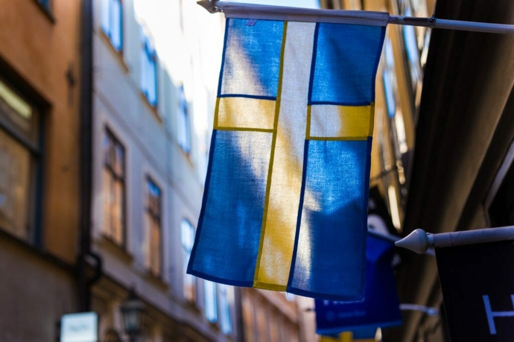 A Swedish flag with a yellow cross on a blue background is mounted on a pole, fluttering in the breeze. It is set against a backdrop of blurred urban buildings under a clear sky.